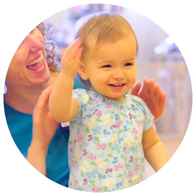 Baby standing smiling with mom sitting behind her smiling