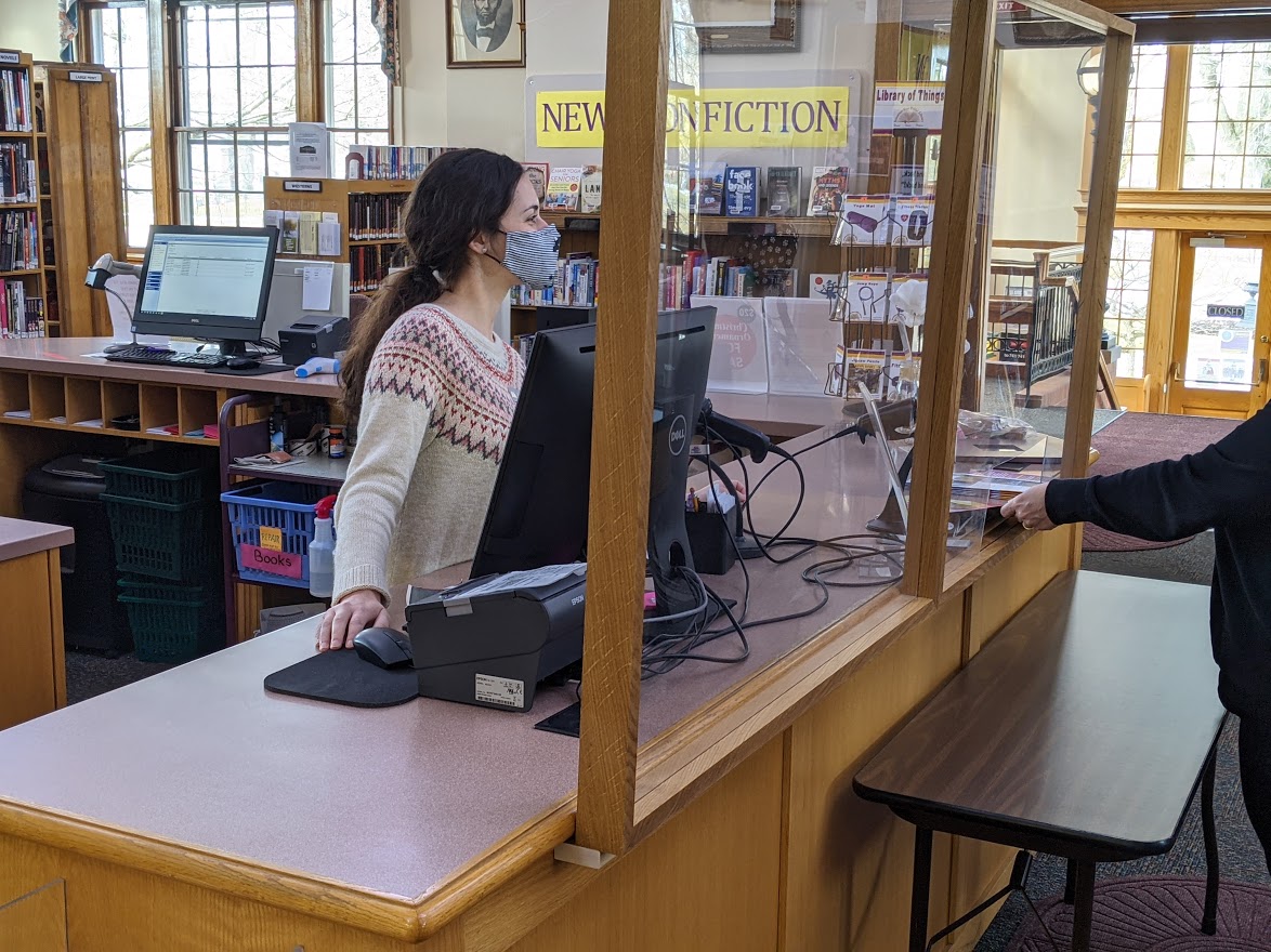 Woman standing at library circulation desk wearing a mask and smiling