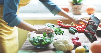 Person wearing an apron with vegetables on a table using hand to tap tablet for recipe