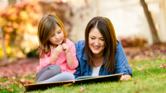 A woman laying on the grass smiling looking at an open book in front of her and a little girl sitting next to her also looking at the book